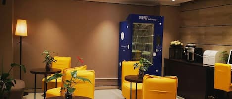 A modern hotel lobby with yellow armchairs, a reception desk, and a blue vending machine.