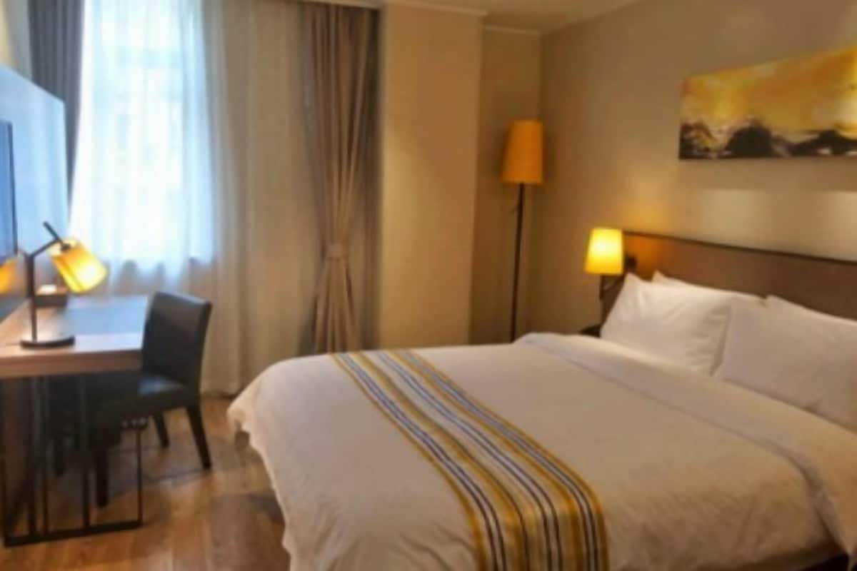 A hotel room with a bed, a desk with a chair, a lamp, and a painting on the wall.