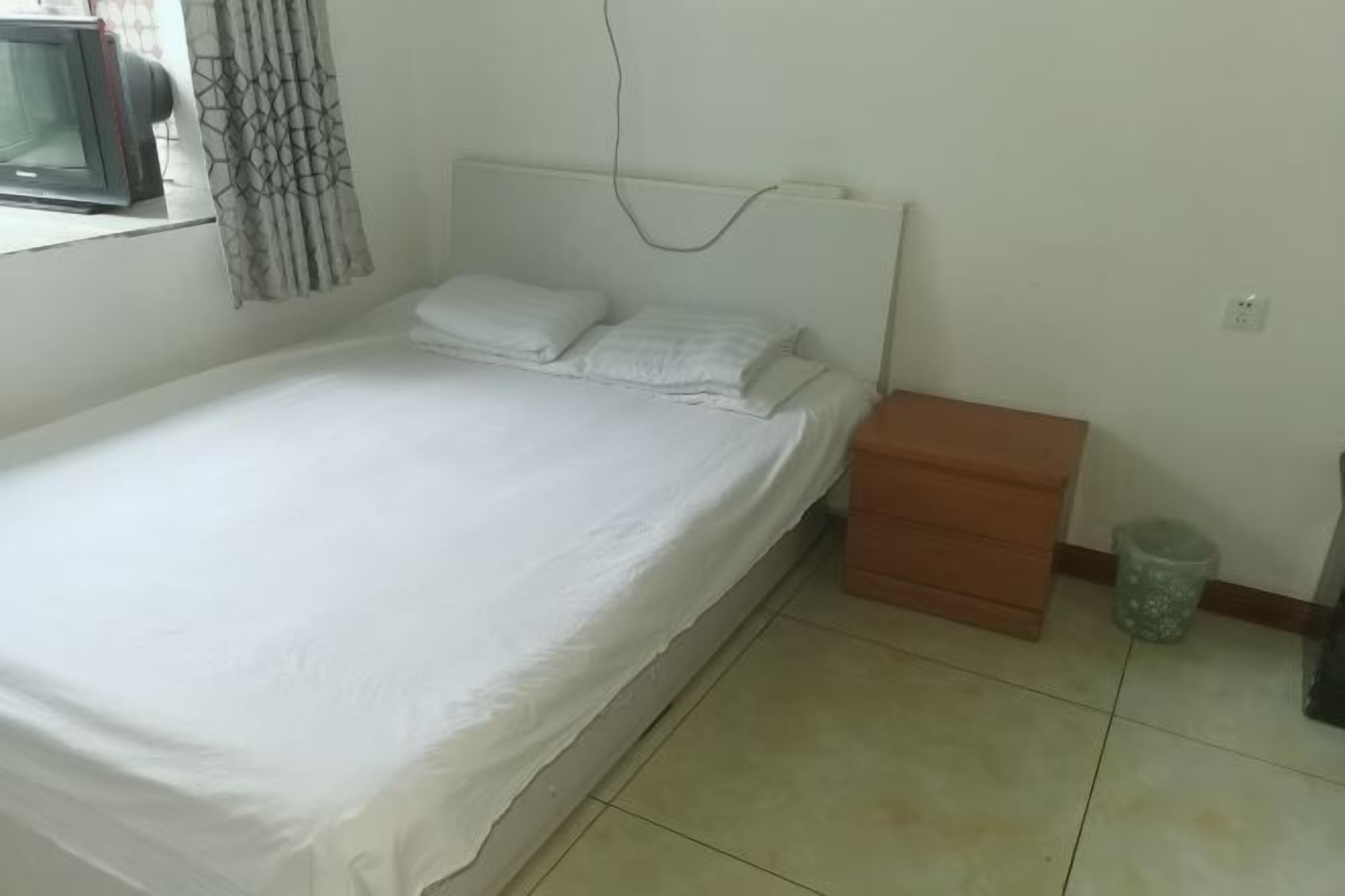 A single bed with white bedding, a nightstand, a trash bin, and a television on a stand.