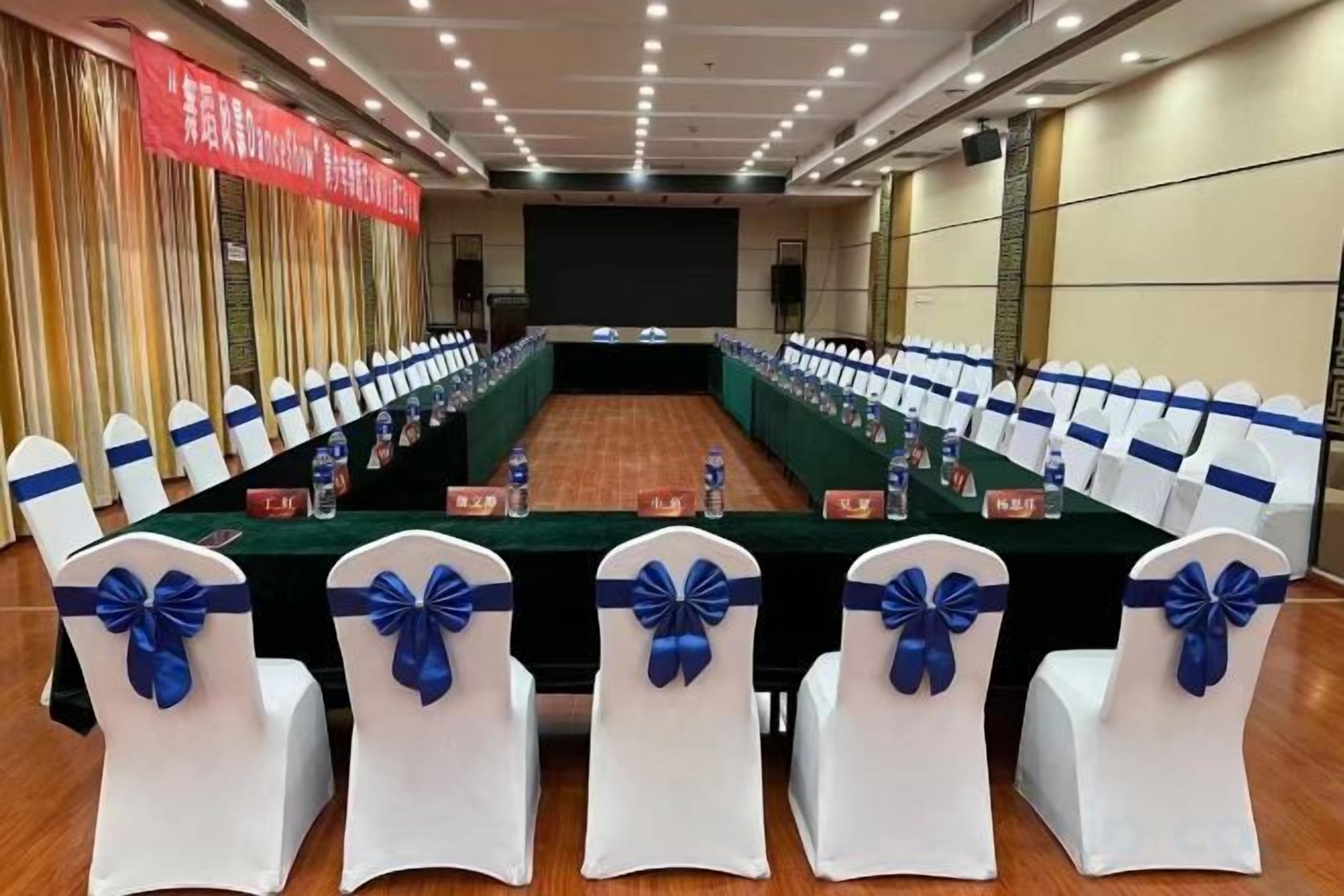 Une salle de conférence avec une longue table, des chaises blanches ornées de rubans bleus et une banderole rouge accrochée au mur.