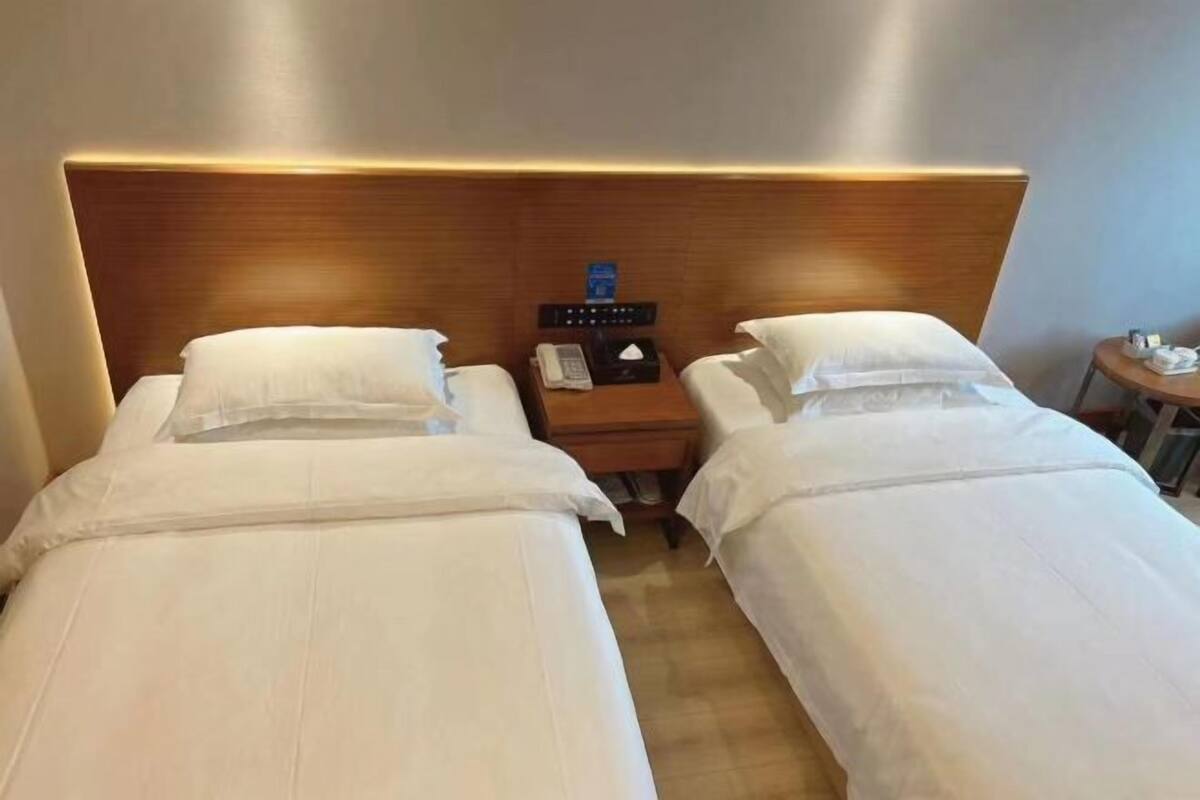 A hotel room with two single beds, a wooden headboard, a bedside table with a phone and tissues, and a small round table with a lamp.