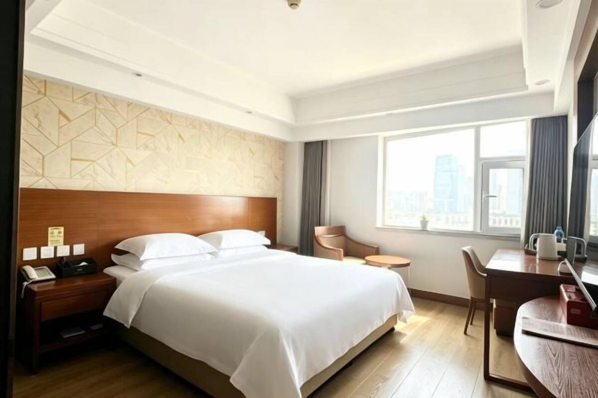 A hotel room with a large bed, a desk, a chair, a nightstand, and a window with a city view.