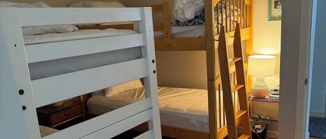 Ein Zimmer mit Etagenbetten, einem kleinen Stuhl mit gemustertem Sitzkissen, einer Holzleiter, einer Lampe und einem Teppich auf dem Boden.