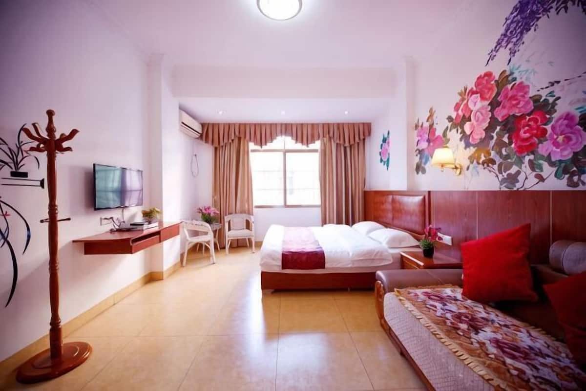 Habitación de hotel con cama, escritorio con silla, televisor, perchero y un ventanal con cortinas.
