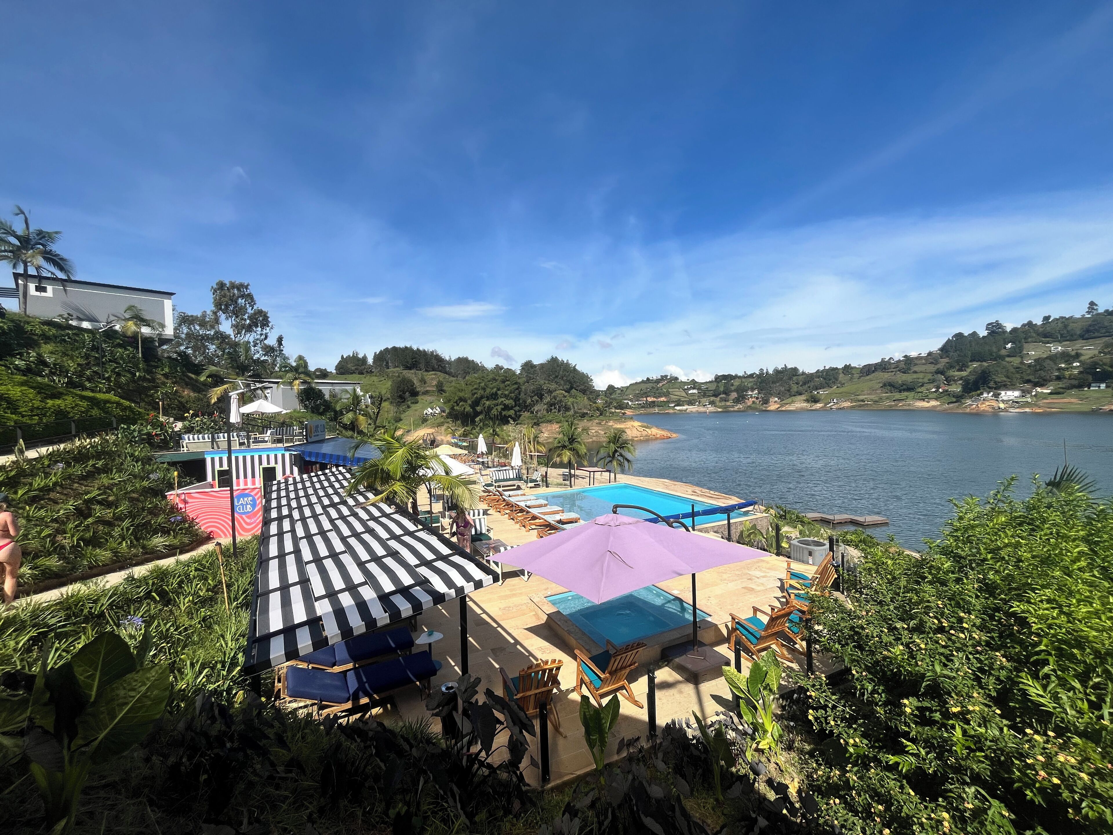 Zona de piscina con sillones reclinables y sombrillas, rodeada de vegetación exuberante y con vistas a un lago.