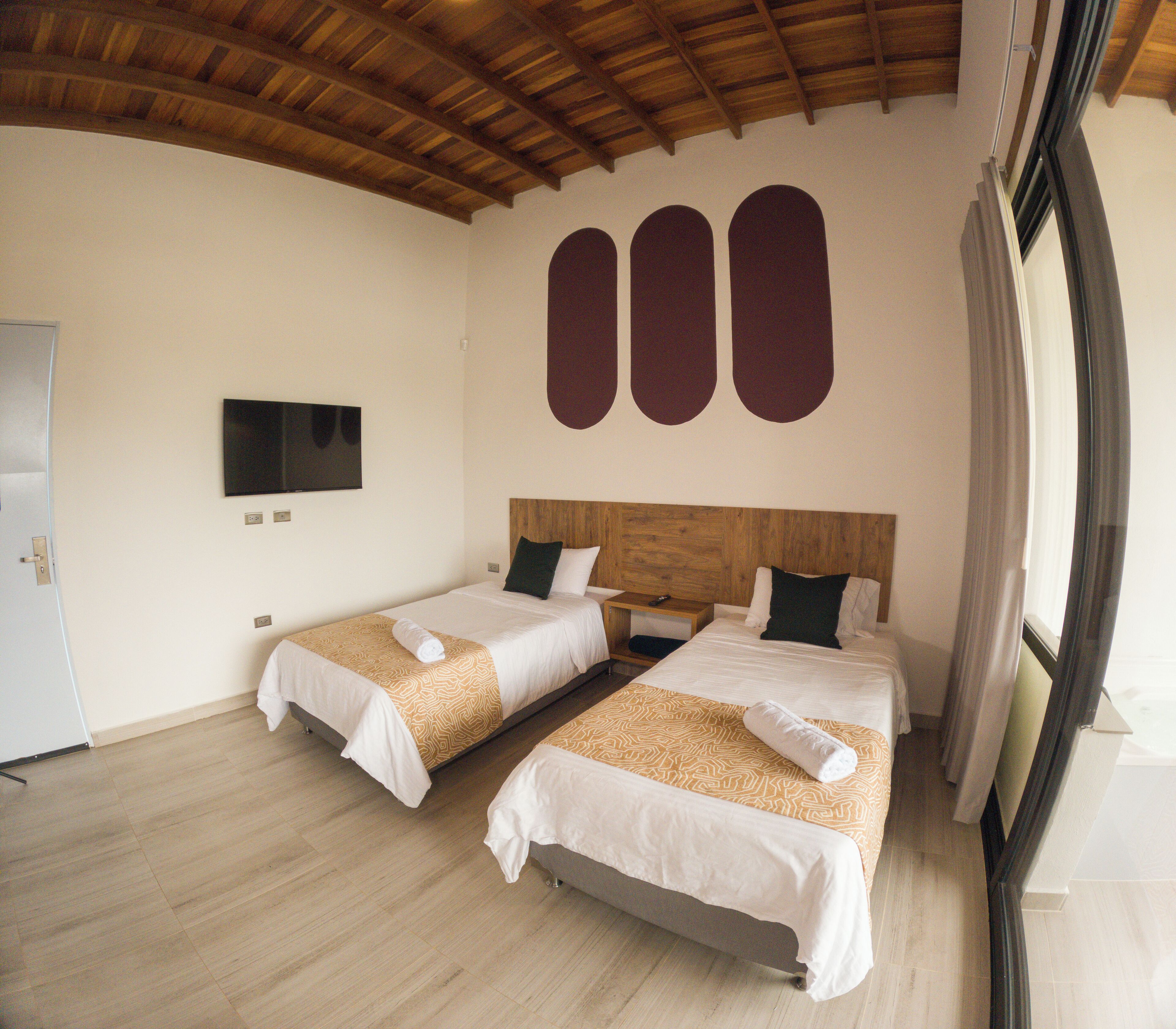 A hotel room with two beds, a wooden ceiling, a TV on the wall, and a window with curtains.