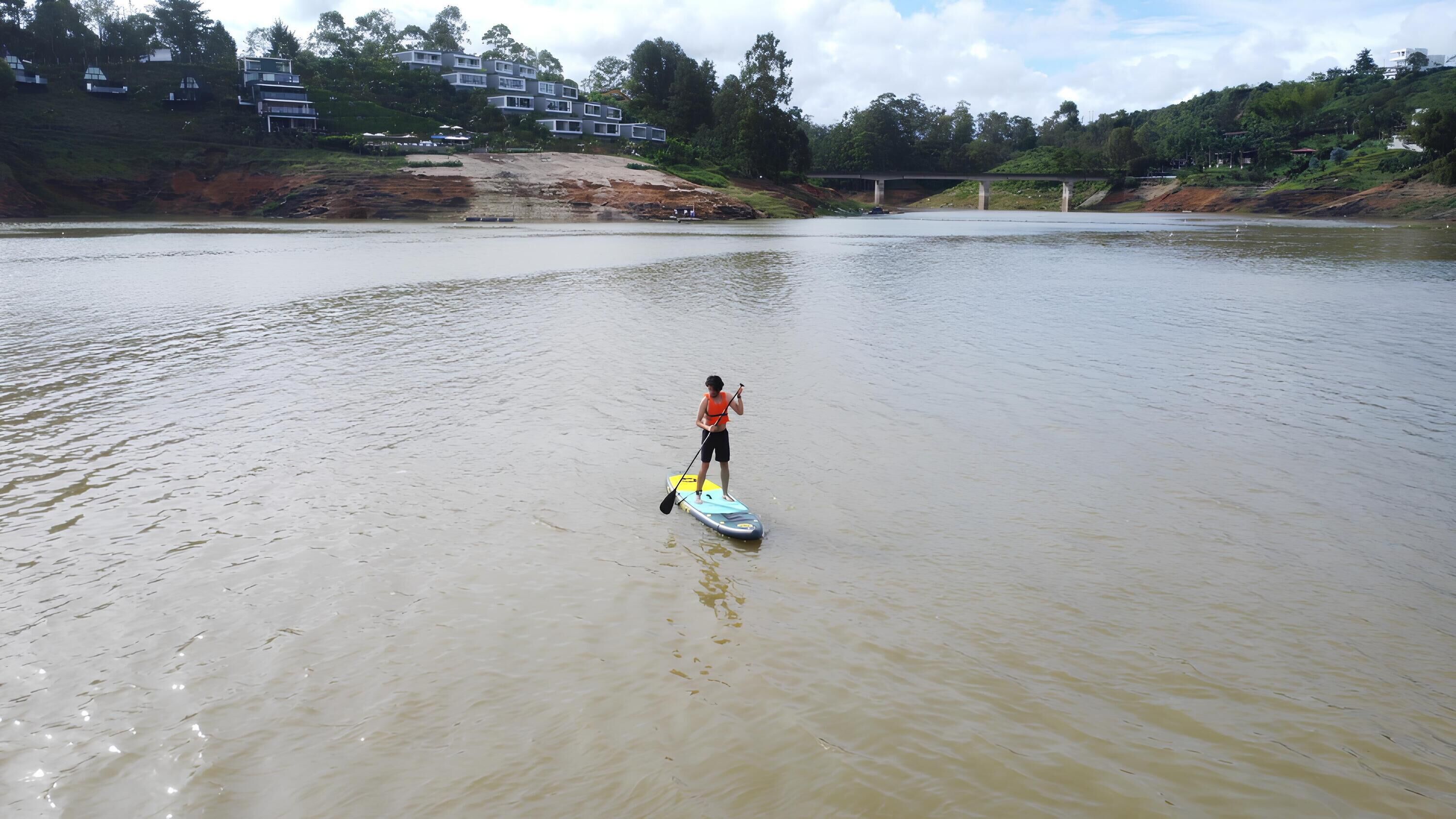 A person paddleboarding on a calm river with a bridge in the background.