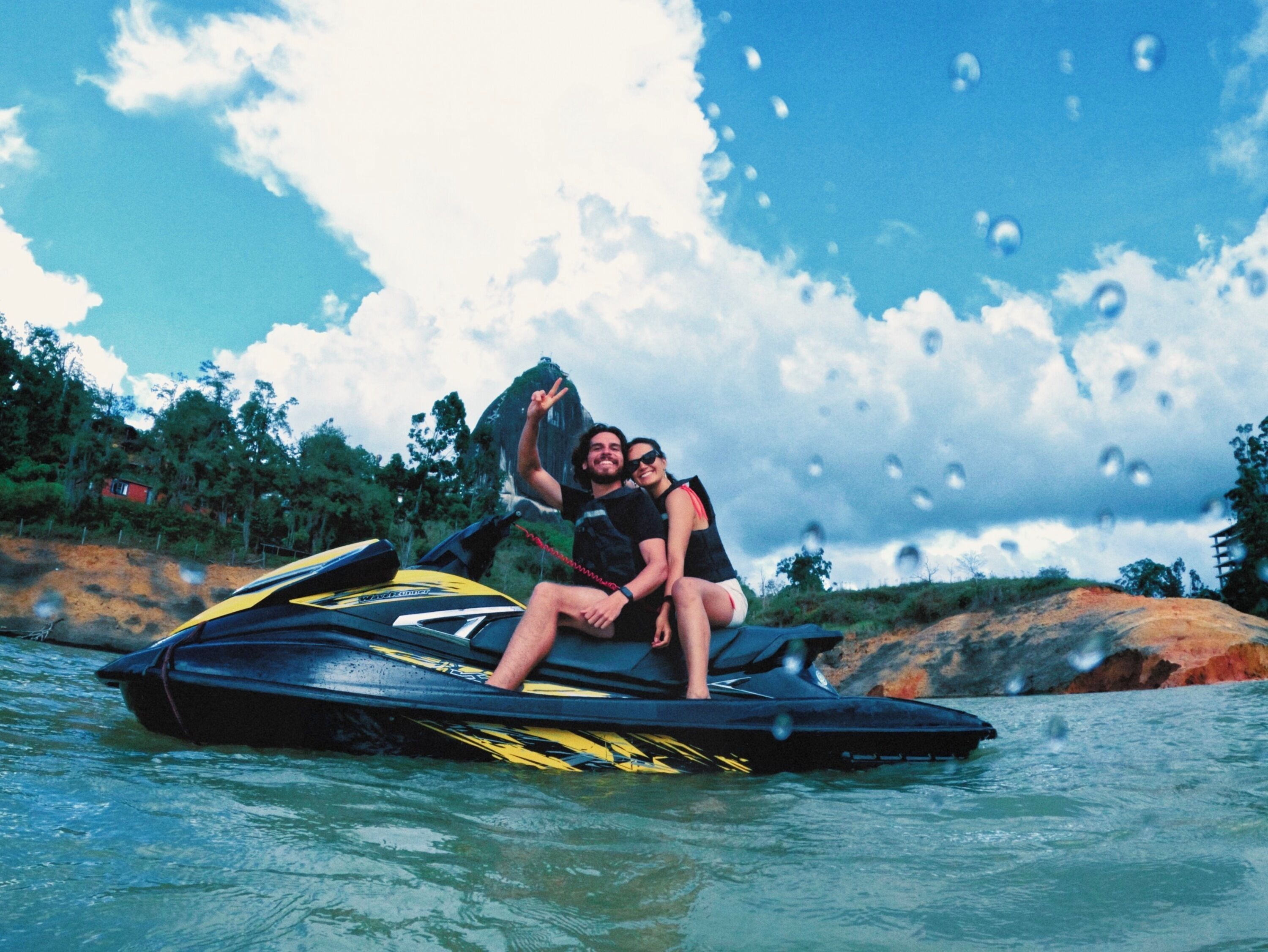 Two people on a jet ski in a body of water with a rocky shoreline and trees in the background.