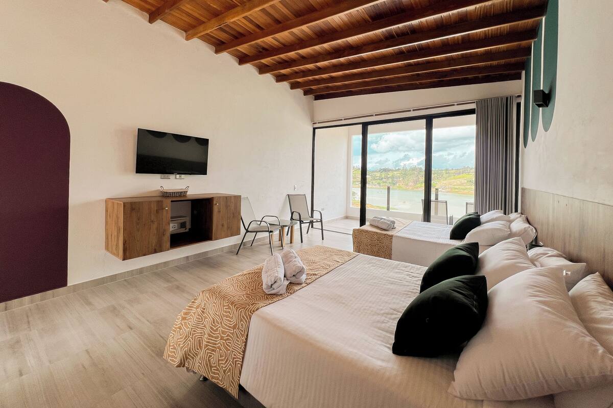 Une chambre moderne avec un plafond en bois, un lit agrémenté de plusieurs coussins, une télévision et une vue sur un lac grâce à de grandes fenêtres.