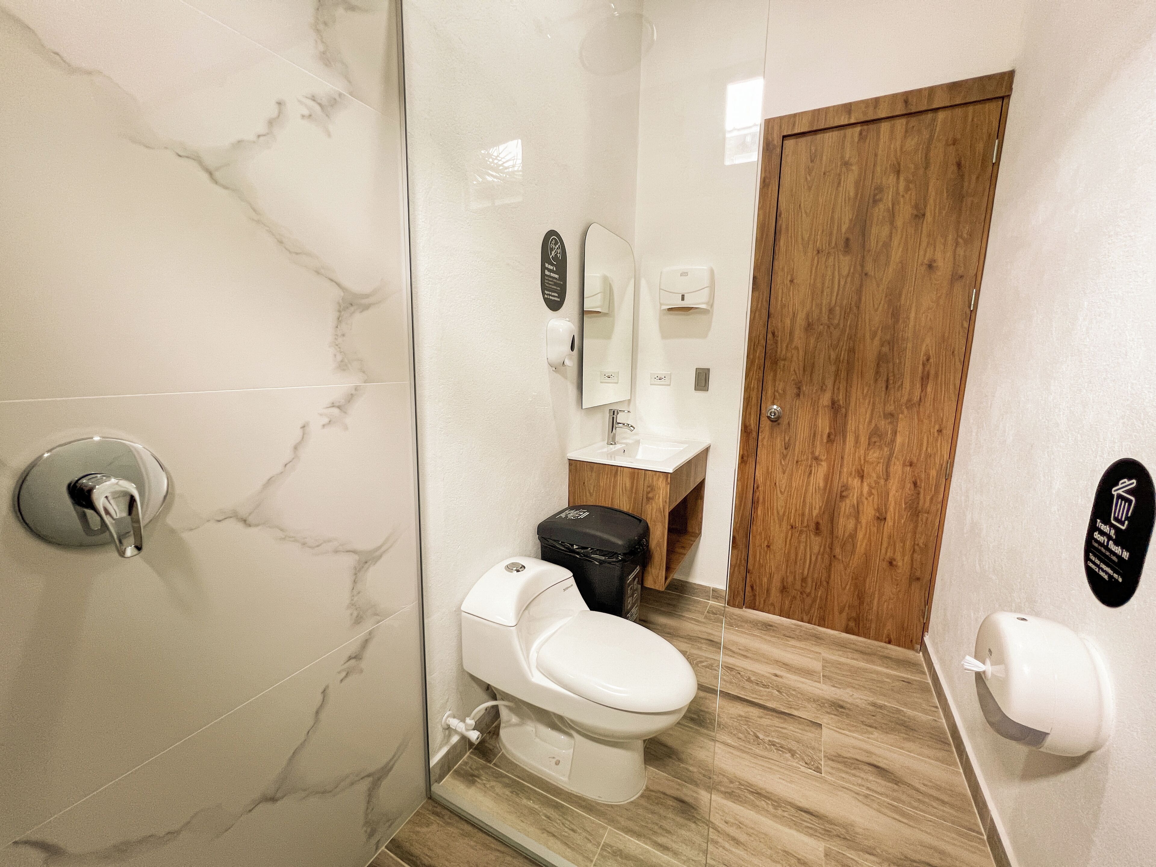 Een badkamer met een marmeren wand, een houten deur, een toilet, een wastafel en een prullenbak.