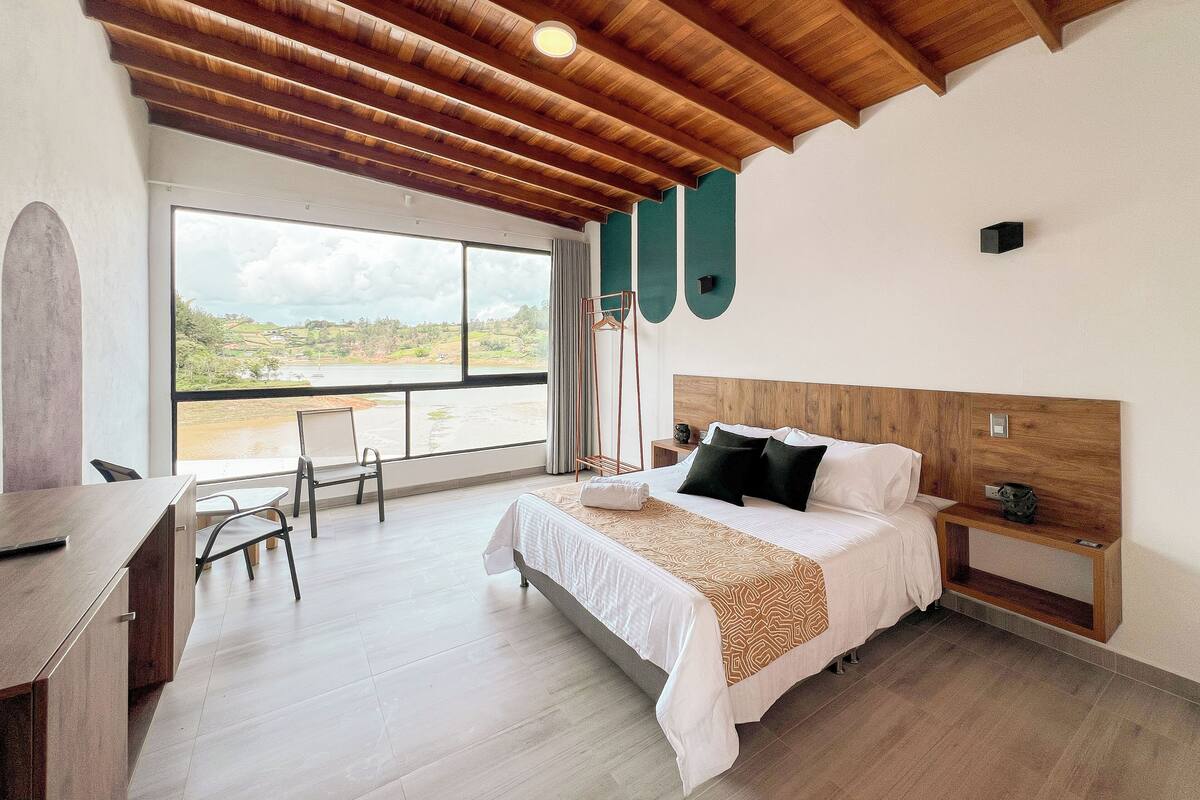 Une chambre avec un plafond en bois, une grande fenêtre donnant sur la campagne, un lit avec une literie blanche et un couvre-lit à motifs, et un bureau avec une chaise.