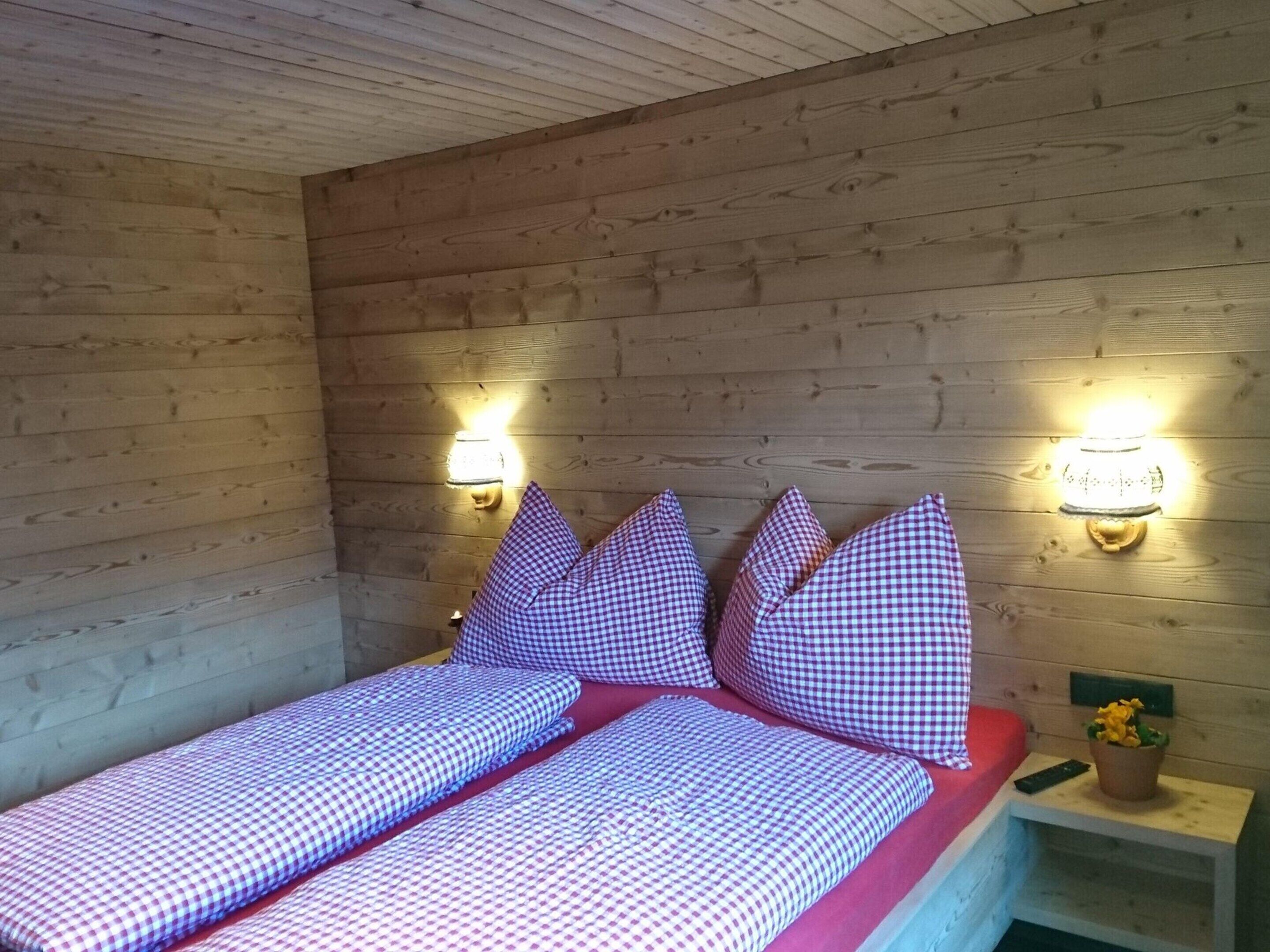 Habitación con paredes de madera, dos camas individuales con ropa de cama cuadriculada, lámparas de pared y una pequeña mesa con una planta.
