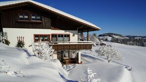 Eine zweistöckige Chalet mit Holzbalkon und einem mit Schnee bedeckten Dach, eingebettet in eine verschneite Landschaft.