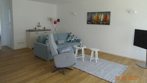 Un salon moderne avec un canapé gris, une petite table basse, un fauteuil gris, un tableau représentant des voiliers accroché au mur et un téléviseur à écran plat.