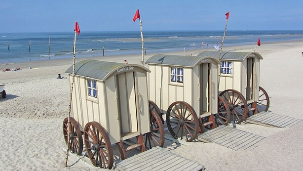 Des cabanes de plage dotées de roues en bois, une plage de sable et l’océan en arrière-plan.