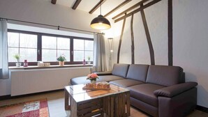 A modern living room with a sectional sofa, a wooden coffee table, a large window with curtains, and a hanging pendant light. - Holiday Apartment Alpakahof Blomberg (Meinerzhagen)