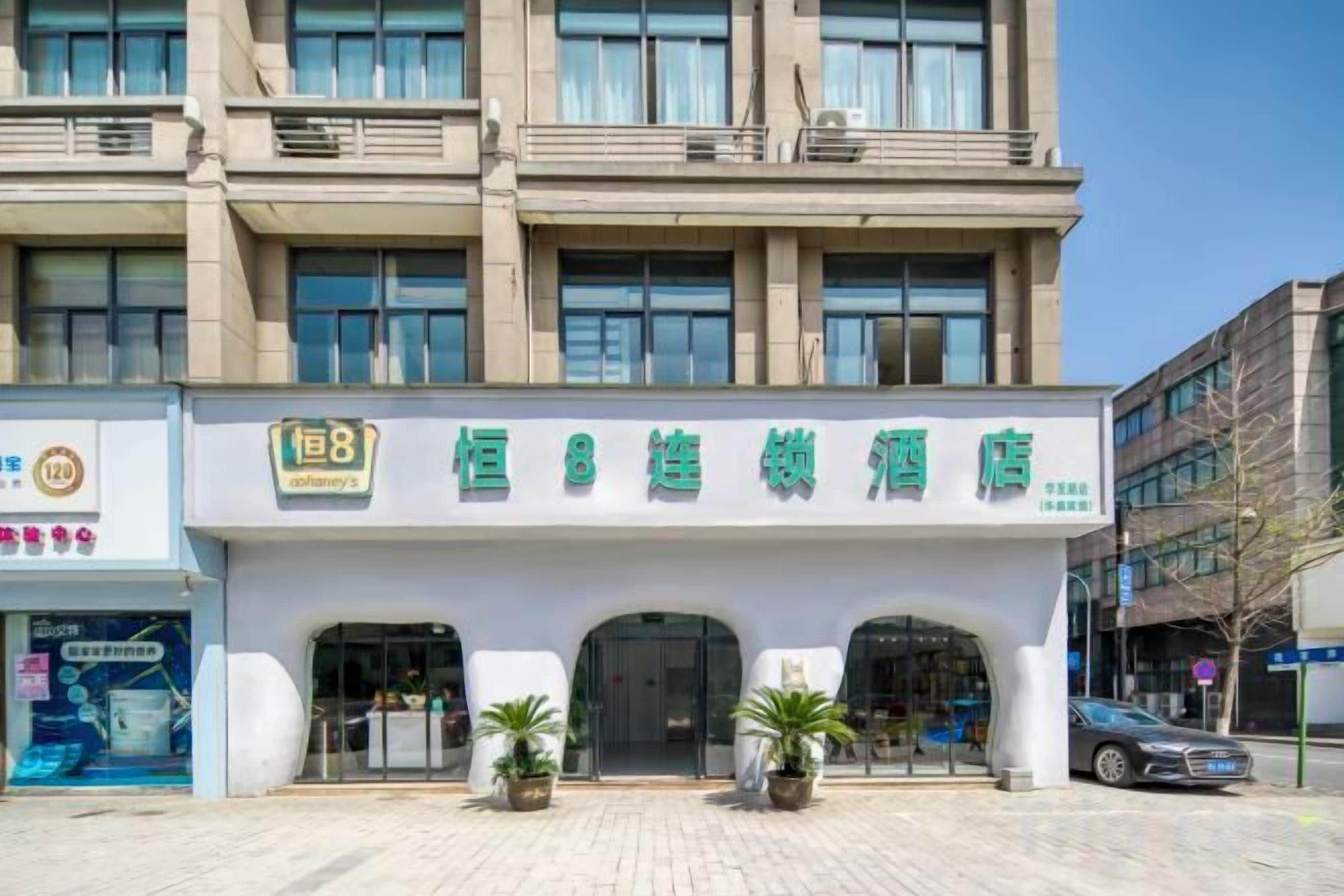 Un hotel con un letrero llamativo, varios pisos y un acceso pavimentado con plantas en macetas.