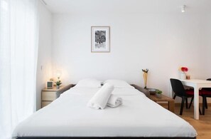 Een netjes opgemaakt bed met witte lakens en twee opgerolde handdoeken, een klein nachtkastje met een lamp, een ingelijst beeld op de muur en een bureau met een stoel en een vaas met bloemen.