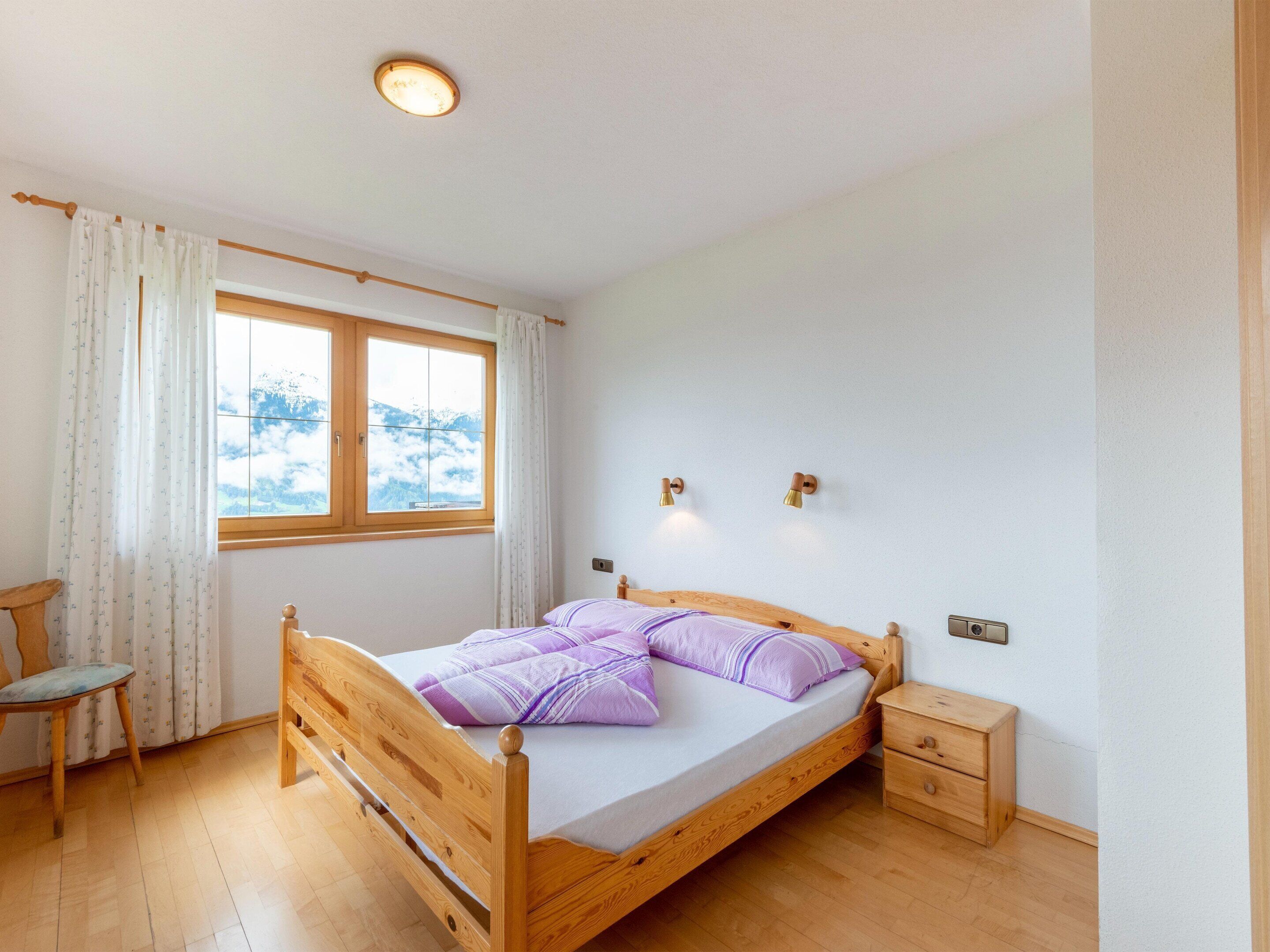 A bedroom with a wooden bed, a nightstand, a chair, and a window with curtains.