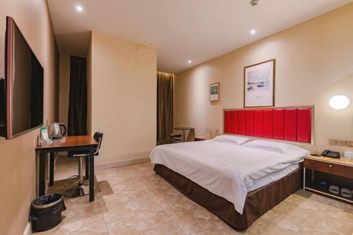 A hotel room with a bed, a desk, a chair, a trash bin, a bedside table with a lamp, and a framed picture on the wall.