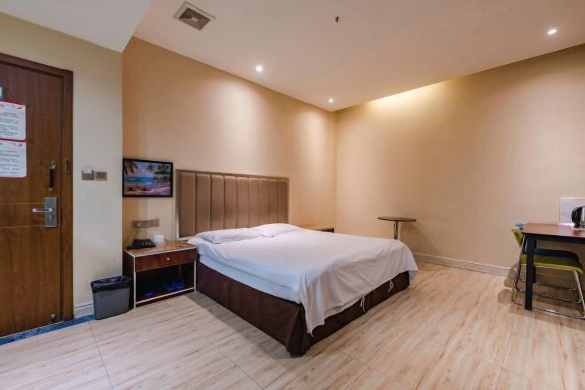 A bedroom with a bed, a desk, a chair, a trash bin, and a wall-mounted picture.