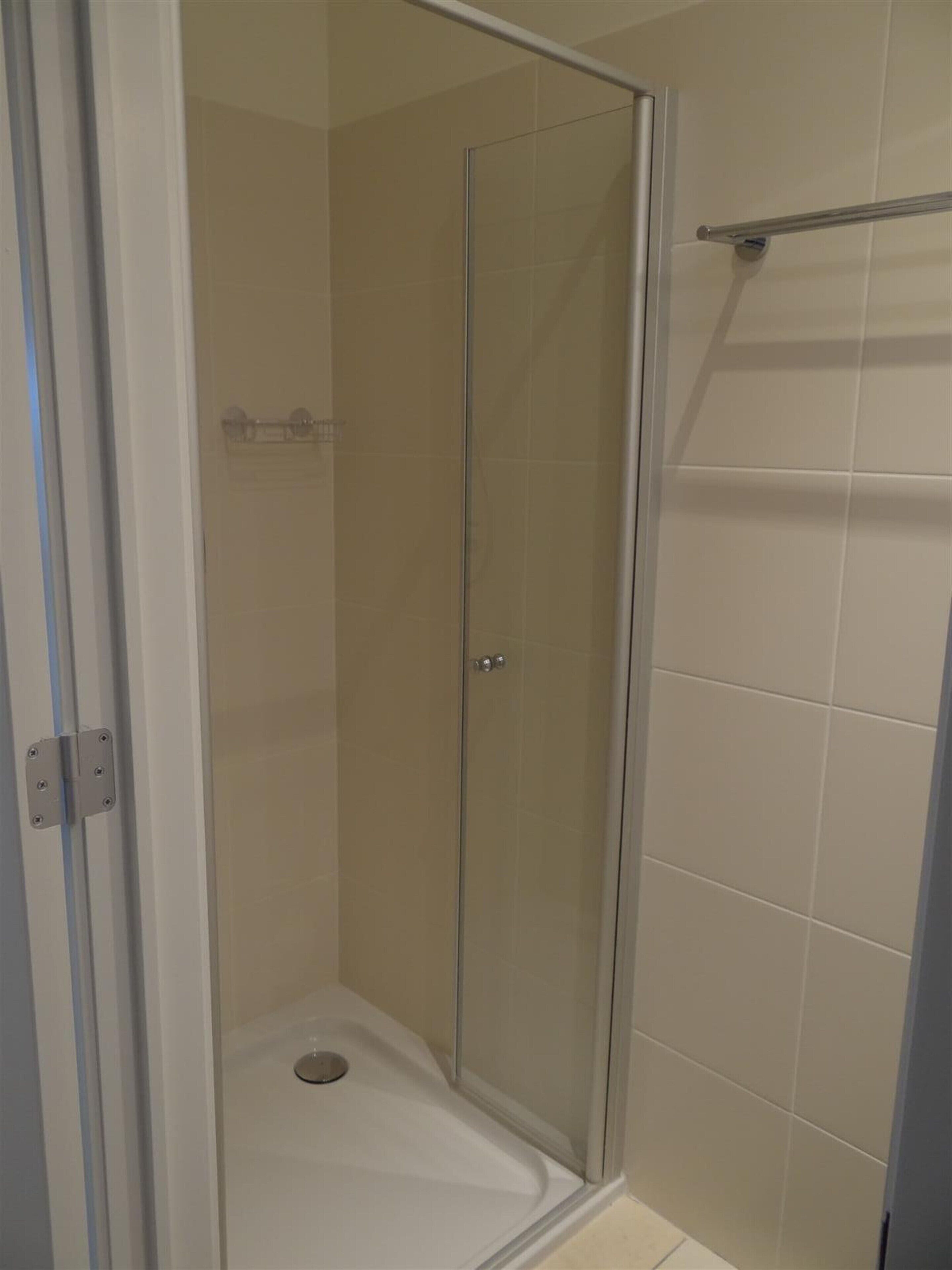 Une cabine de douche fermée par des parois en verre, avec un sol blanc, un porte-serviettes en métal et des murs recouverts de carreaux beiges.