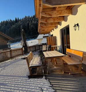 Eine hölzerne Terrasse im Freien mit Bänken und Tischen, umgeben von einem Holzzaun, mit Schnee auf dem Boden und einem Wald im Hintergrund.