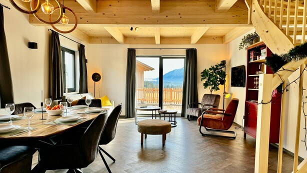 Een eethoek met een houten tafel voor vier personen, een bank en een stoel. Er is een groot raam met uitzicht op een houten terras en bergen.