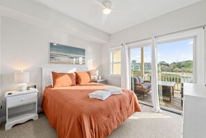 A bedroom with a large bed, a nightstand, a lamp, a painting, and a view of the outside through a sliding glass door. - Aviation Themed Townhome w/ Waterpark & Game Room - Pet Friendly (Kill Devil Hills)