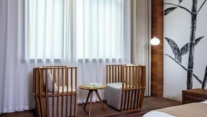 Ein Zimmer mit zwei Holzstühlen, einem kleinen runden Tisch, einem Bett und einer Wand mit einem Gemälde einer Bambuspflanze.