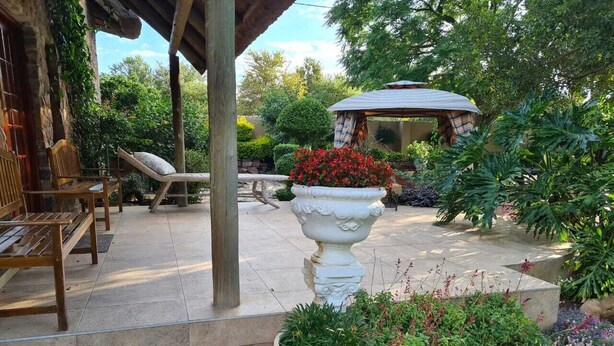 Un espace patio agrémenté d’un banc en pierre, d’une chaise en bois, d’un vase blanc orné de fleurs rouges et d’une pergola.