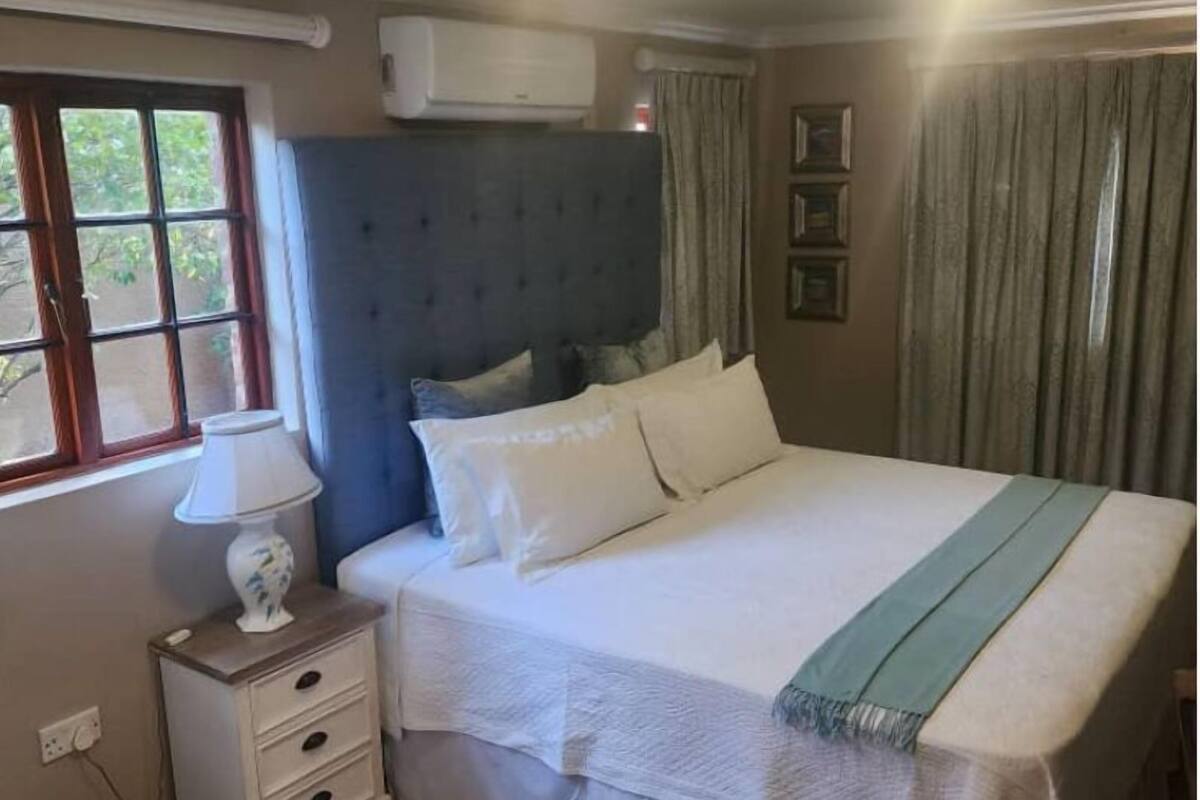 A bedroom with a bed, a bedside table with a lamp, a window, and an air conditioning unit.