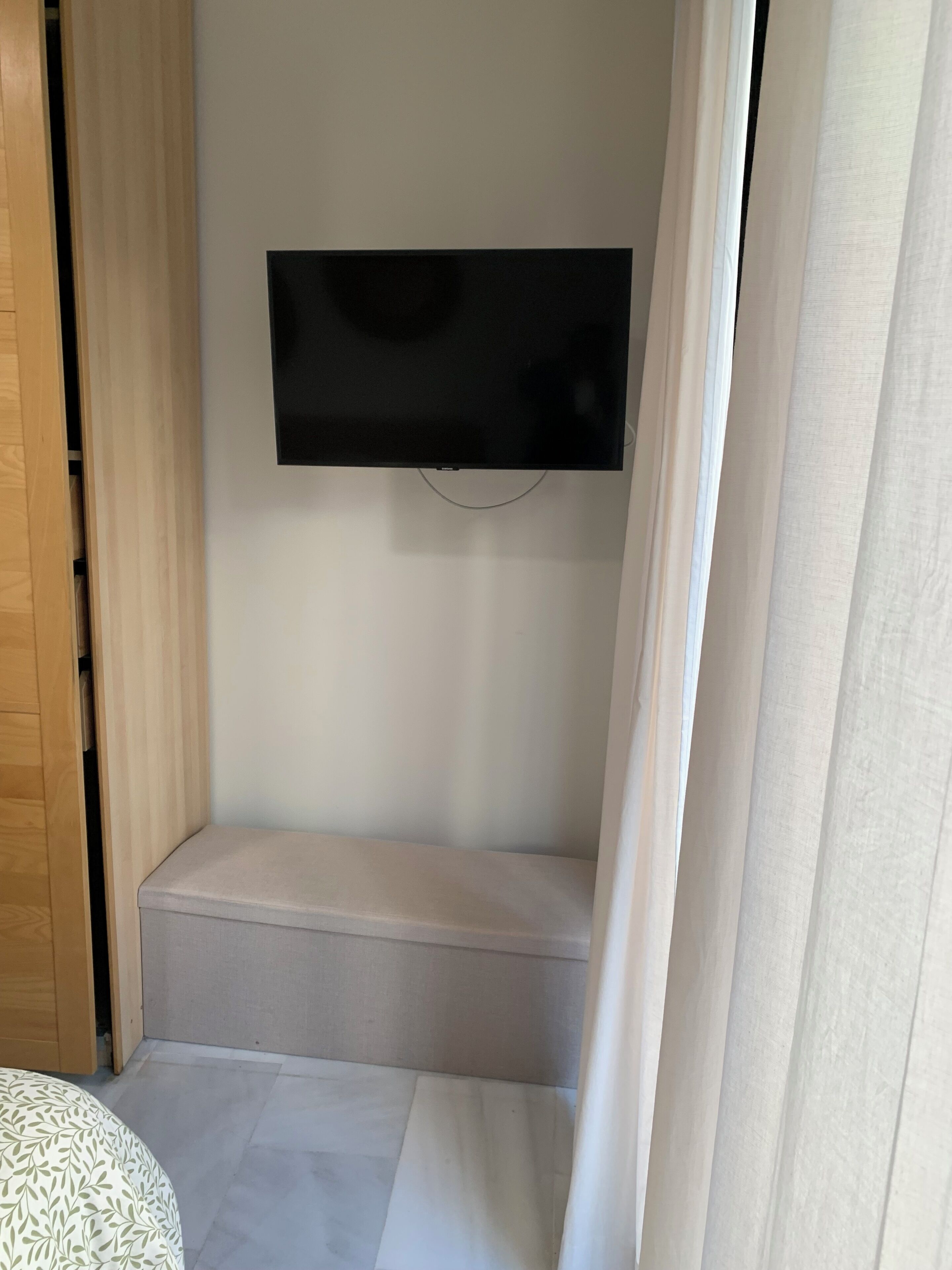 Una habitación pequeña y estrecha con un televisor de pantalla plana en la pared, un armario de madera y una colcha estampada.