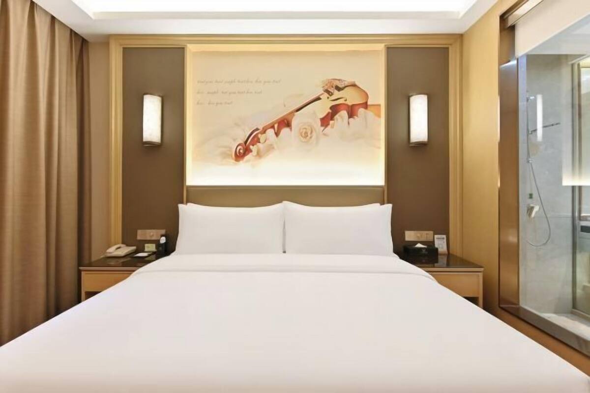 A hotel room with a large bed, a nightstand with a phone, a wall-mounted lamp, and a glass shower enclosure.