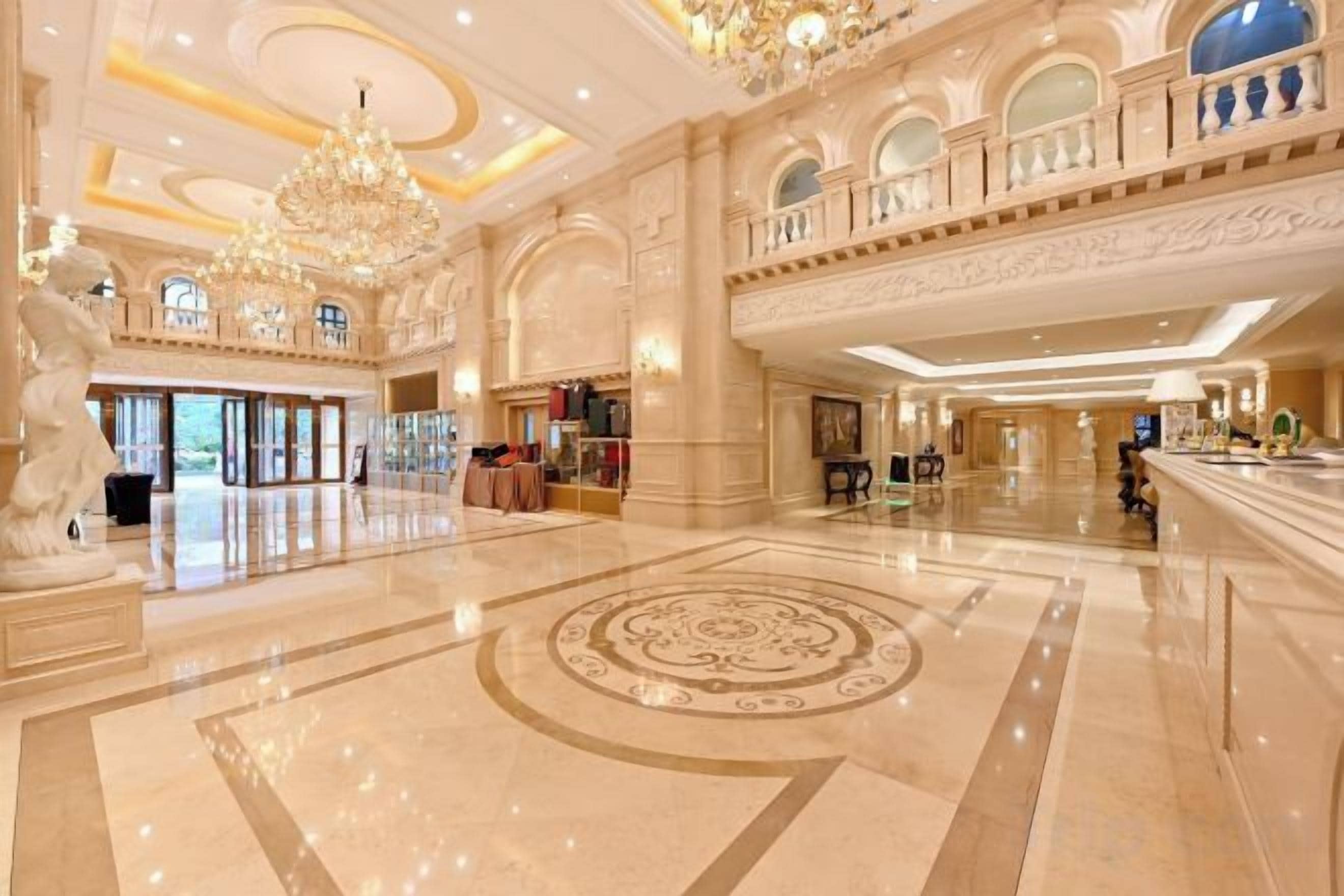 A spacious lobby with a grand chandelier, a central circular floor design, and a balcony overlooking the area.