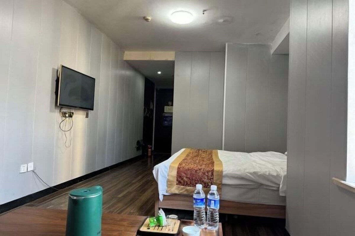 A hotel room with a bed, a flat-screen TV mounted on the wall, a wooden floor, and a small table with water bottles and a green container.
