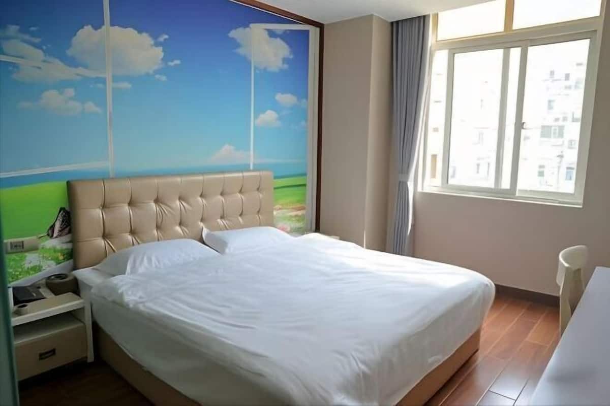 Un dormitorio con una cama grande, una mesita de noche, una silla, un ventanal con vistas a edificios y un mural en la pared.