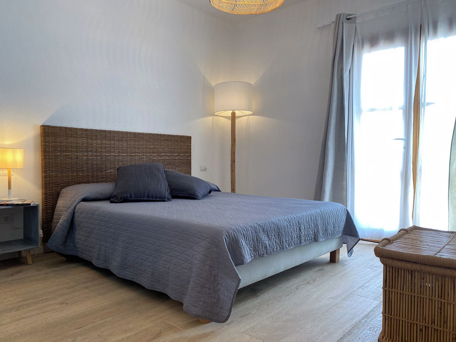 A bedroom with a bed, a nightstand, a lamp, a wicker chair, and a window with curtains.
