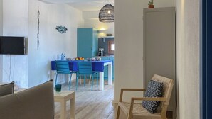 A modern living room with a TV, a dining area with blue chairs, a kitchen in the background, and a cozy seating area with a patterned cushion. - Apt. Azure, central, parking, top for 6 (San Pantaleo)