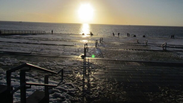 A beach with a pier extending into the water, a few people walking, and the sun setting over the horizon.