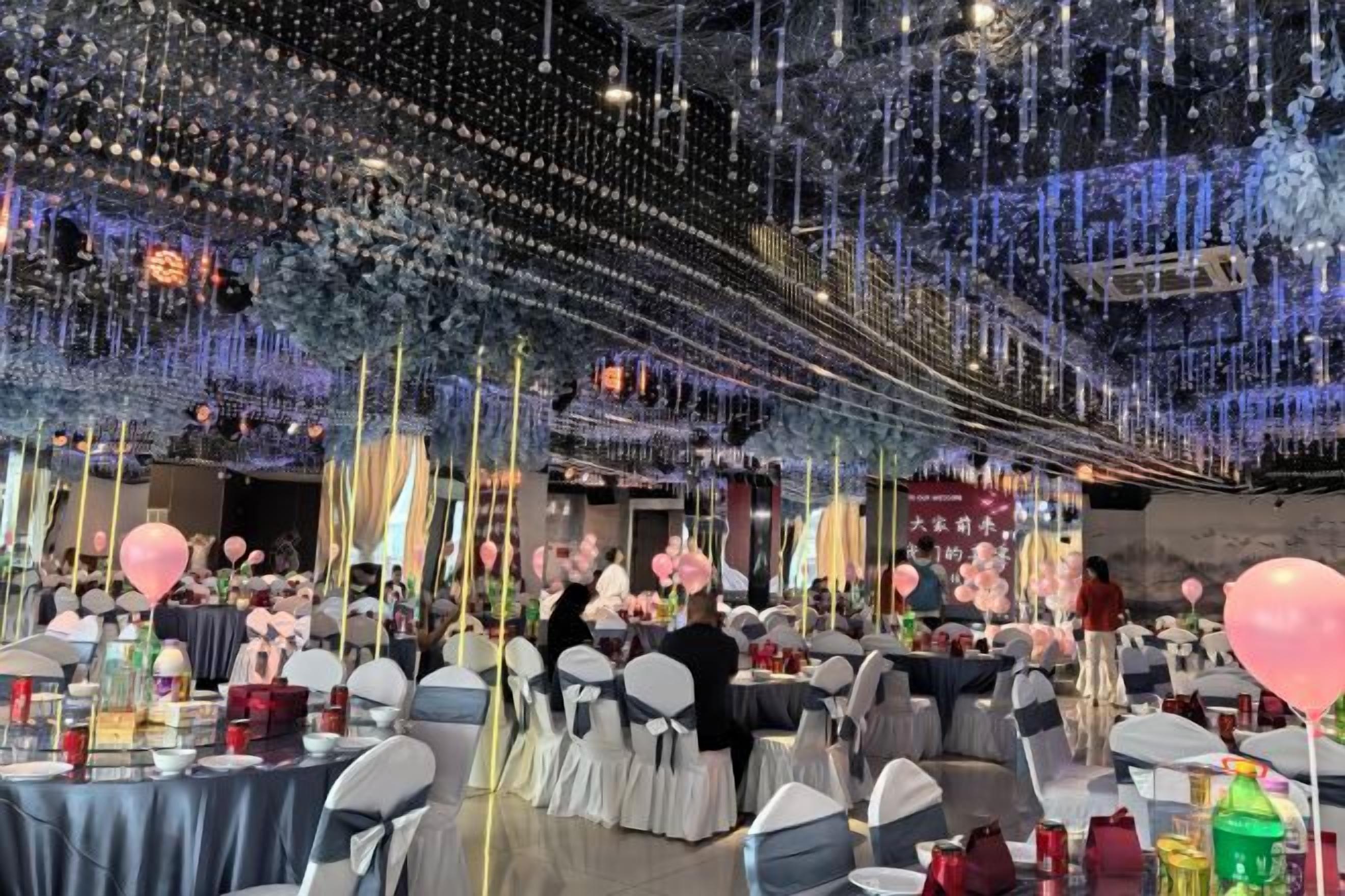 Un salón de banquetes con mesas dispuestas para un evento formal, decorado con luces colgantes y arreglos florales.