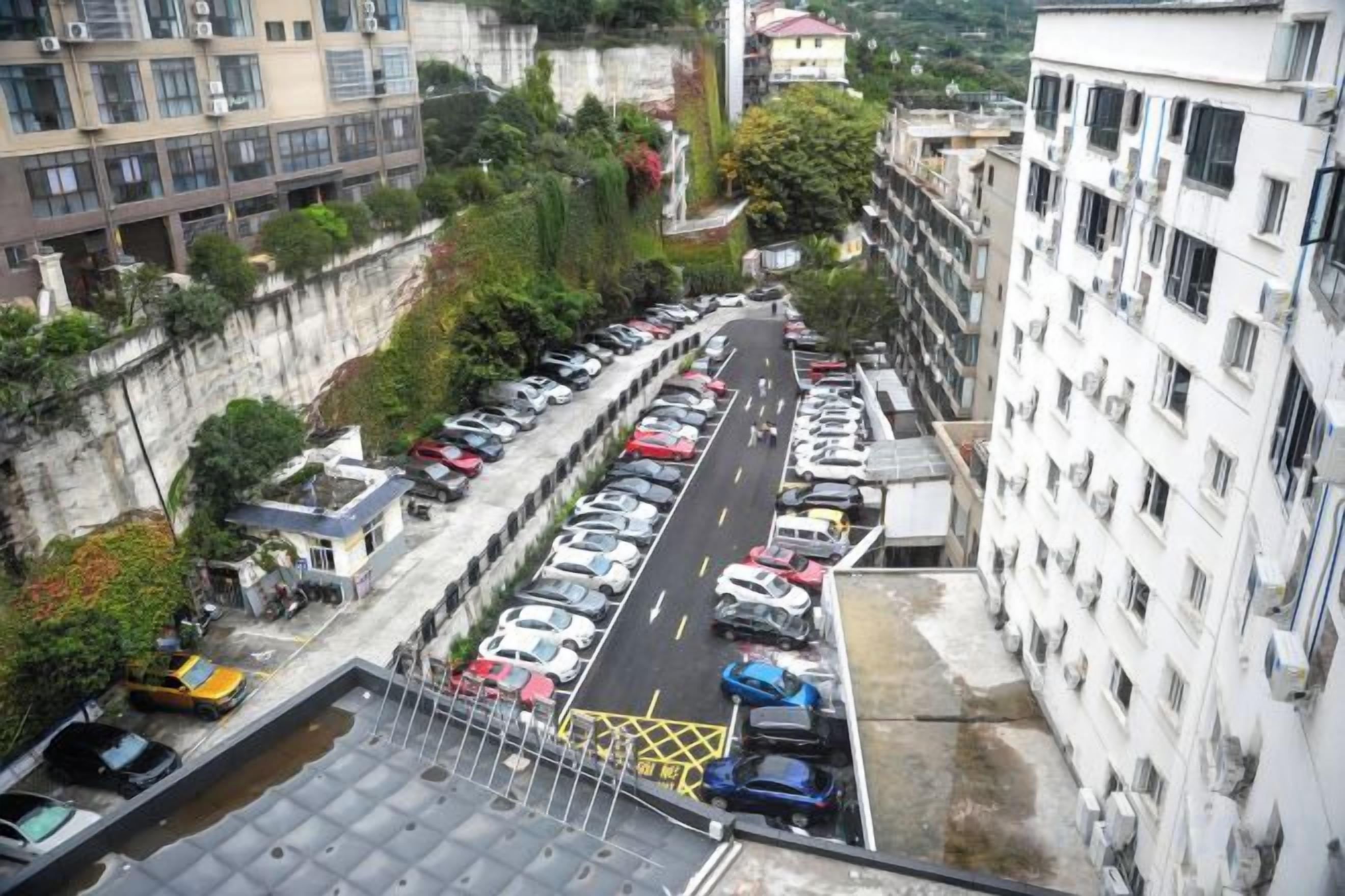 A multi-story parking garage with rows of parked cars, adjacent buildings, and a small green area.