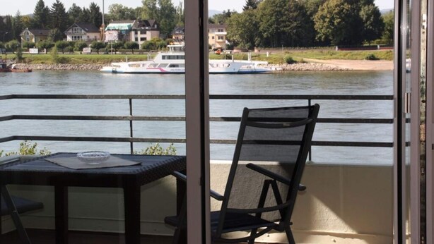 Un balcon avec une table et une chaise, donnant sur une rivière où l’on aperçoit des bateaux et des maisons au loin.