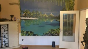 A room with a large tropical mural, a white door, and a small desk with a lamp and a bottle.