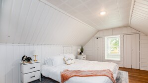 A bedroom with a sloped ceiling, a bed with white bedding, a bedside table with a lamp, a nightstand with a clock, and a window with a view of greenery.