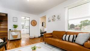 A modern living room with a brown leather sofa, a white coffee table with a plant, wooden flooring, a kitchen in the background, and a large wall clock.