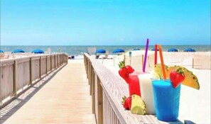 Une promenade en bois menant à une plage où l’on trouve des parasols et des boissons.