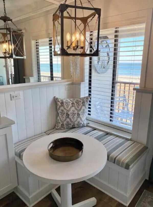 A cozy seating area with a round table, white paneled walls, and a view of the beach.