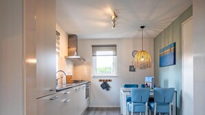 A modern kitchen with white cabinets, a built-in oven, and a window. There is a dining area with blue chairs and a table. - Holiday Home Near Dunes With Wellness Garden (Den Burg)