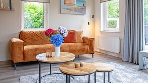 A modern living room with a brown sofa, two round wooden tables, and a large window with a view of greenery. - Holiday Home Near Dunes With Wellness Garden (Den Burg)